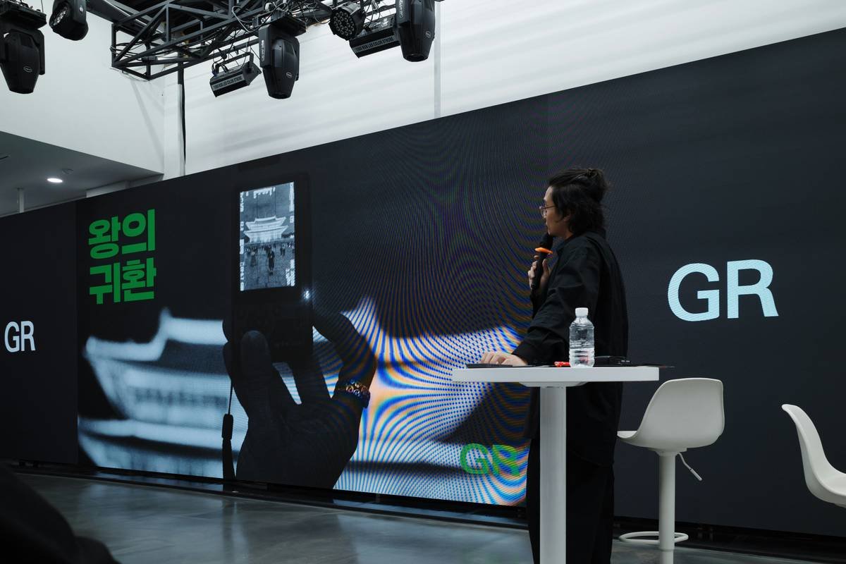 A person speaks at a presentation with a large screen.